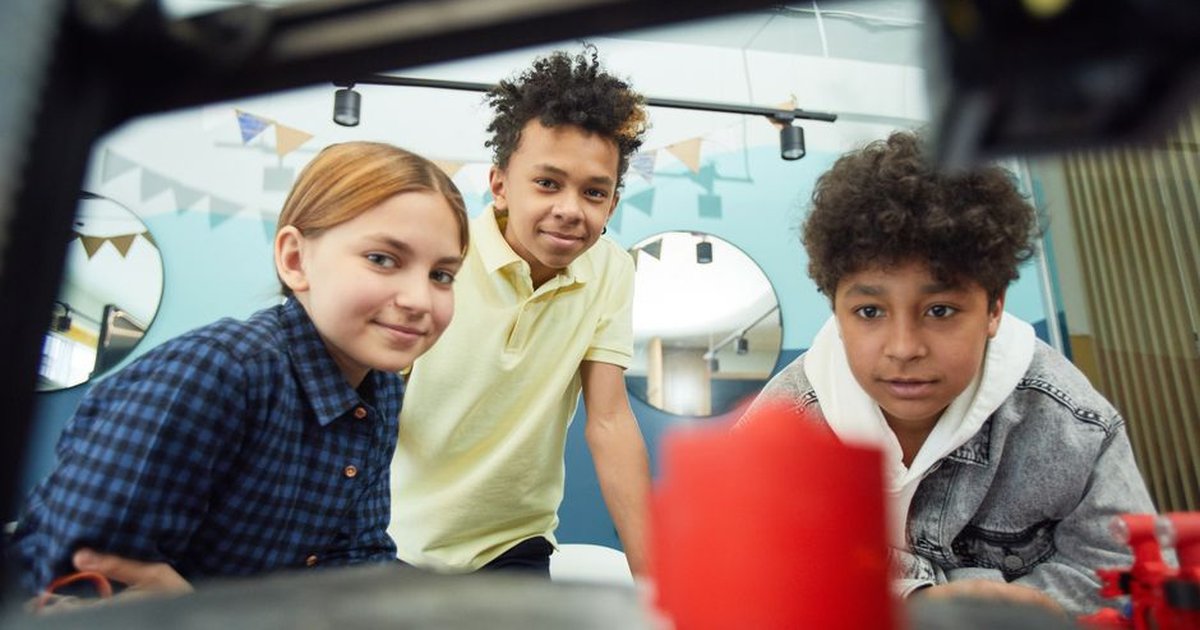 Low angle of cheerful group of multiracial children sitting together and observing work of 3D printer in light workshop