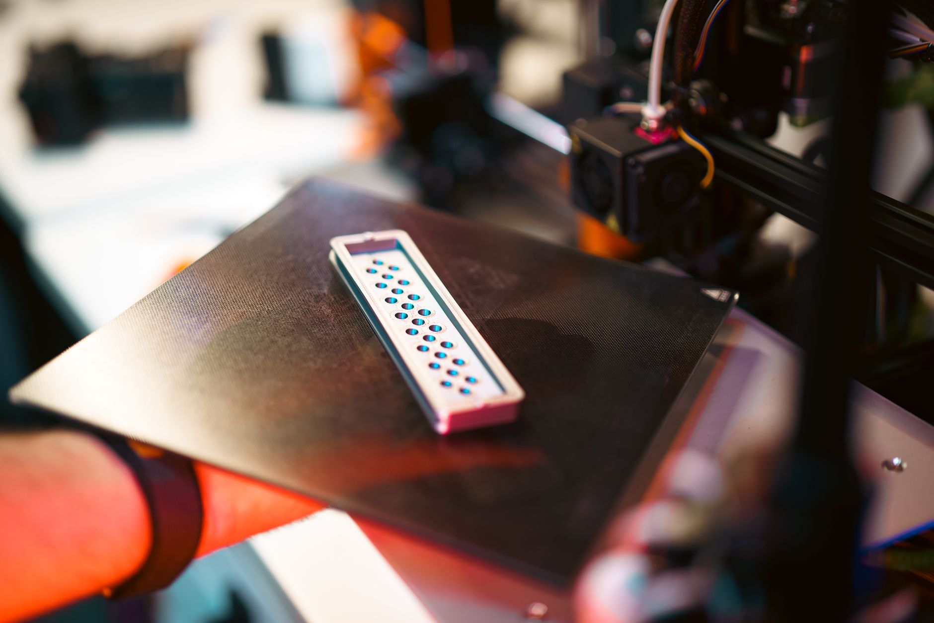 A hand carefully removes an orange 3D printed object from a printer bed using a spatula indoors.